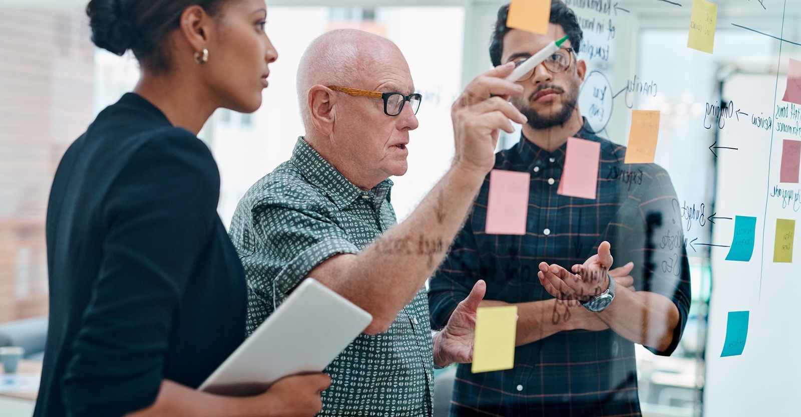 A small team collaborating around a glass wall covered in colorful sticky notes, with one person pointing and writing while others observe and discuss.